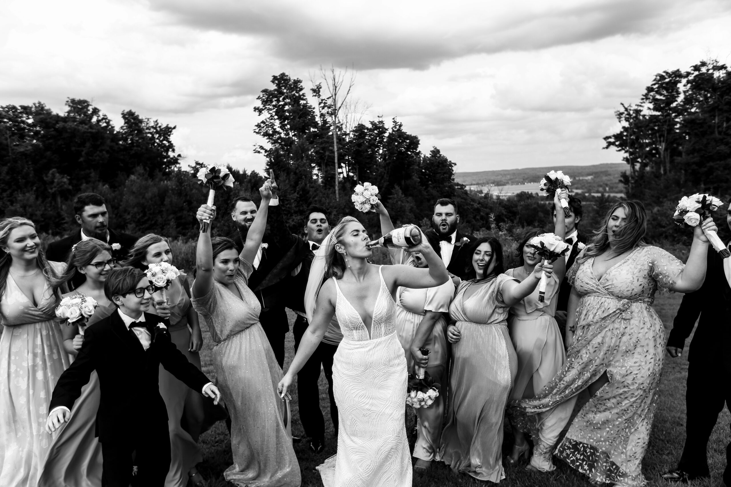 A bride drinking from a champagne bottle while the wedding party cheers around her.